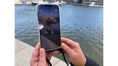 Eine Person Hält ein Handy auf der die Bremerhaven Guide App vor dem Zoo Am Meer gezeigt wird mit einem Eisbären auf dem Bildschirm